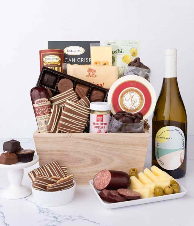 Wooden crate filled with gourmet snacks and a bottle of white wine, including summer sausage, creamy cheeses, chocolate-covered grahams and almonds, assorted chocolates, wafers, crisps, mustard, and olives, styled with plates of cheese and sausage.