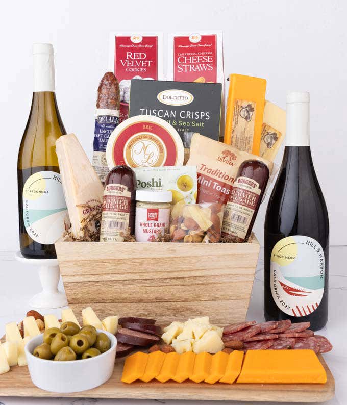 Wooden basket filled with 4 cheeses, 3 sausages, crackers, cookies, nuts, mustard, olives and crisps alongside 2 bottles of wine - Pinot Noir & Chardonnay.