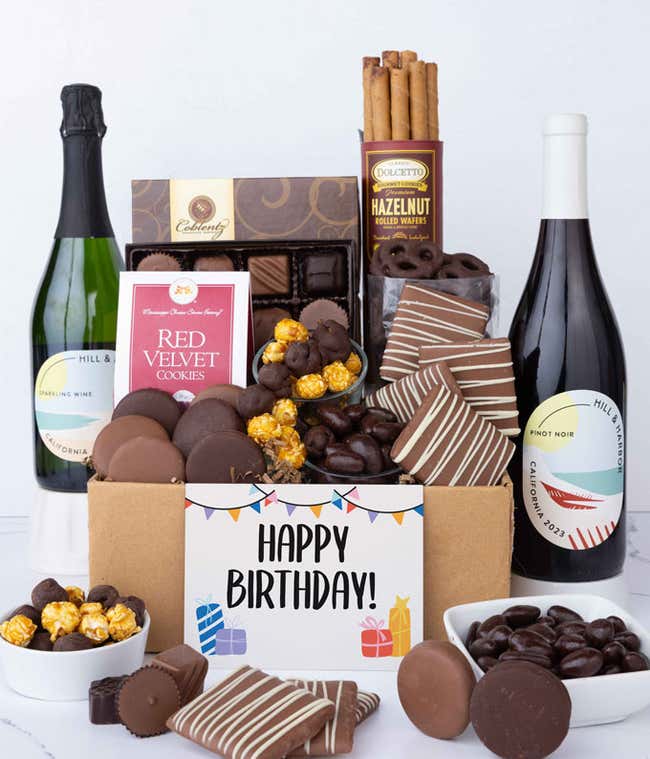 A gift box overflowing with treats including an assortment of chocolate-covered items like oreoes, almonds graham crackers and pretzels along with red velvet cookies, hazelnut wafers, an 8 piece box of chocolates, 2 bottles of wine and a Birthday card.