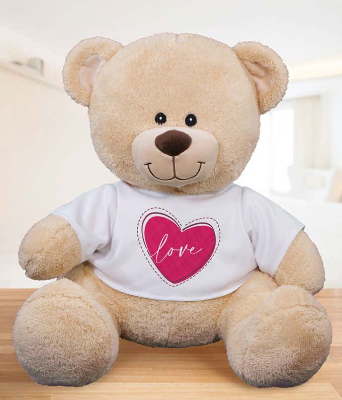 Light tan plush teddy bear sitting on a wooden surface, wearing a white T-shirt with a pink heart design and the word love printed in the center.